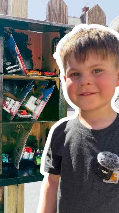 A young boy is standing next to a green toy box that has cars in it. He has short, light-brown hair and is wearing a grey T-shirt.