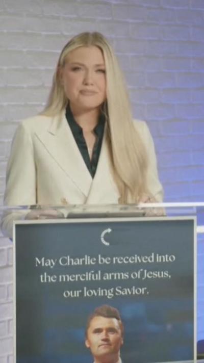 Erika Kirk standing at a podium in a white suit. A sign reads 'May Charlie be received into the merciful arms of Jesus, our loving saviour'