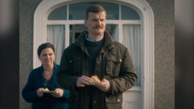 A woman looks in disgust at a moustached man who is in front of her. He is holding a plate of sandwiches