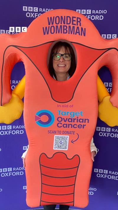 Lis Hale has medium length dark hair and glasses. She is wearing a costume that looks like a pair of ovaries, and has 'Target Ovarian Cancer' written on the front. Behind her is a purple wall with 'BBC Radio Oxford' on it in white writing.