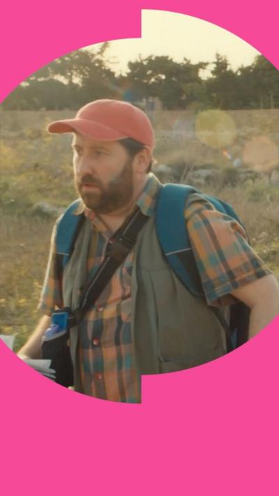 Man wears orange hat, checked shirt and rucksack, and walks along outside in sunshine 