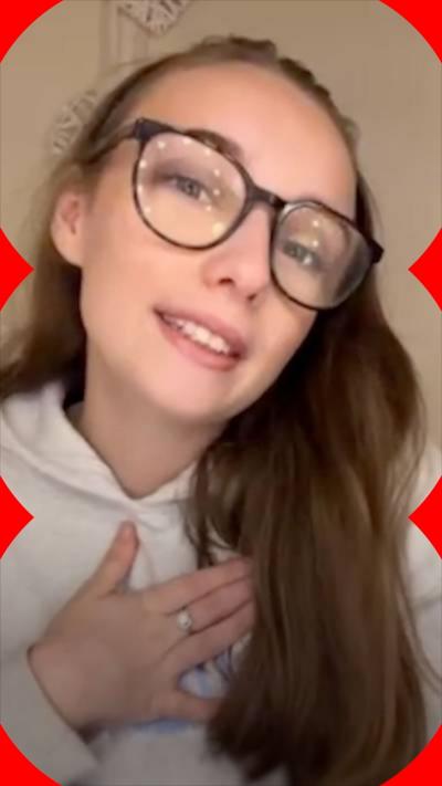 A teenage girl with long brown hair and glasses, holds her hand over her chest. She wears a grey hoodie 