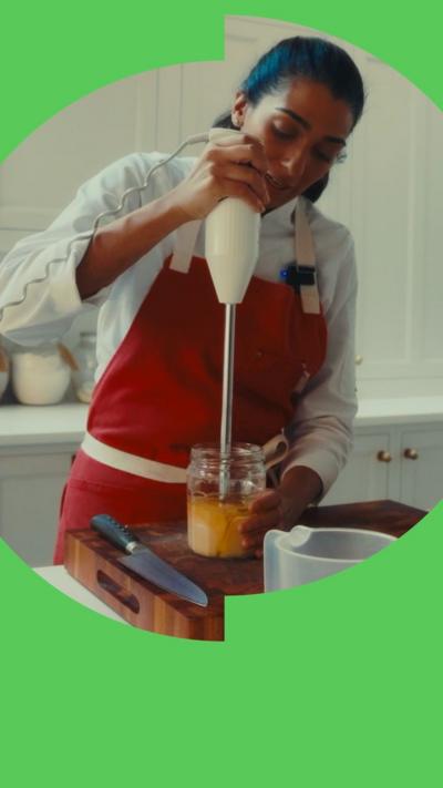 A woman with long, dark hair wearing a red apron over a white shirt blends liquid in a glass jar.