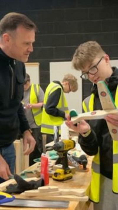 A man in black top and blue jeans helping a young man with glasses with some woodwork work