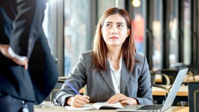 Young woman sat at work, looks up at someone wearing a suit with a hand on their hip