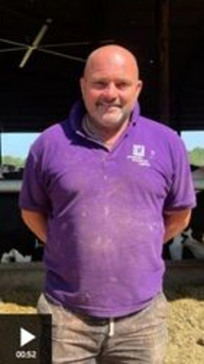A man stands smiling into the camera. He is bald and is wearing a purple polo shirt and brown trousers.