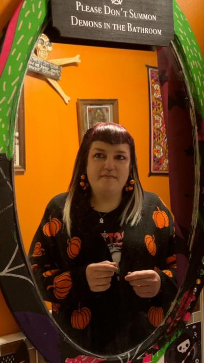 A woman in a black cardigan with pumpkin designs looks into a mirror. A sign says 'Please don't summon demons in the bathroom'