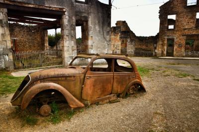 A rusted old car with no wheels surrounded by derelict buildings