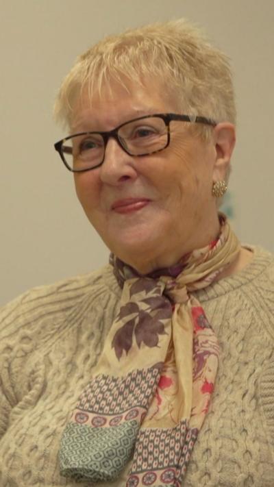 A woman with short, white hair and glasses looks slightly off camera. She has a cream woolly jumper on and a colourful scarf around her neck