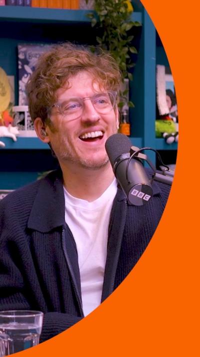 A man in white T-shirt under a blue zip top sits behind a microphone. He is laughing