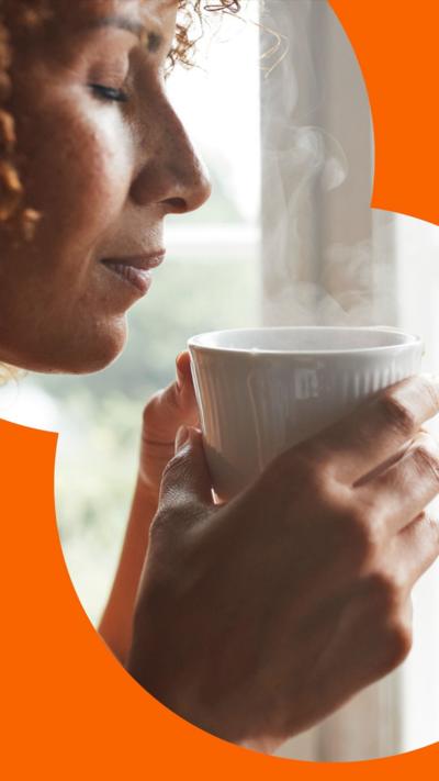 A woman closes her eyes as she holds a cup of coffee next to a window