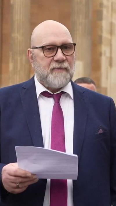 A bald man with glasses, wearing a blue suit, white shirt and purple tie reads a statement outside court flanked by family and friends