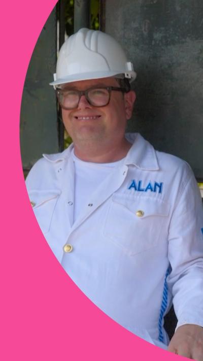 Alan Carr wearing a white helmet and a white boiler suit sitting in a bus stop smiling