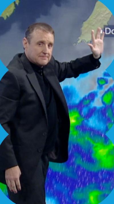 Comedian Peter Kay has short dark hair. He is wearing a black suit and polo neck. He is standing in front of a weather presentation screen which shows a rainfall map. His left hand is raised gesturing to the graphics behind him.