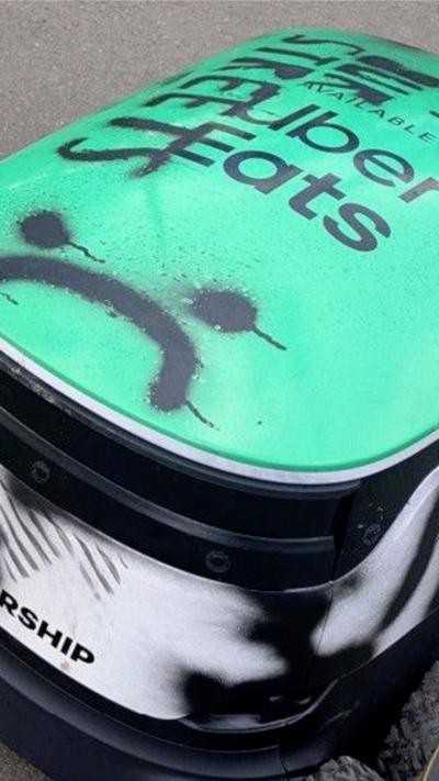 A close up shot of a small white and green robot. On top the words Uber Eats have been obscured by a stencil saying Our Streets and a sad face 