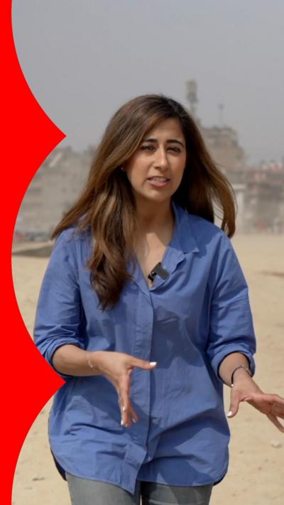 A female reporter with long brown hair walks towards the camera with her hands out. She wears a blue blouse and stands amidst a backdrop of a pothole-filled road.