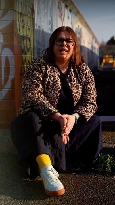 Woman sat near wall in black clothes and leopard print coat.