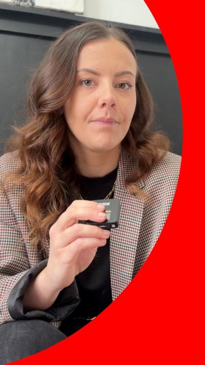 A woman with long brown wavy hair looks into the camera. She is holding a clip-on mic and wearing a jacket with a repeated pattern of lots of small, differently coloured, diamond shapes.