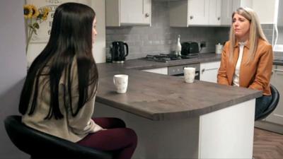 Leanne Lucas sits across from reporter Judith Moritz. They are in a kitchen and both have white mugs in front of them. Leanne's long black hair is down her back and she wears a grey top. Judith wears a dark orange jacket.