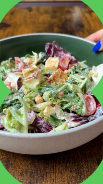 A bowl is shown being placed on a wooden table. It is full of green leaves, tomatoes, chickpeas and halloumi cubes covered in a tzatziki dressing.