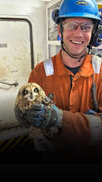 Oil rig worker Sam Cooke wearing orange visibility shirt and blue helmet holding owl Oli
