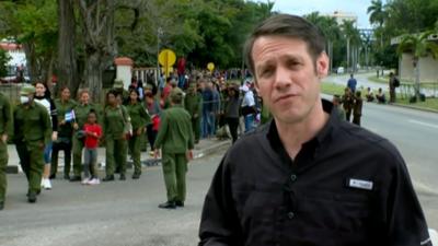 BBC correspondent Will Grant on Cuban street with army personal in the backgroud.