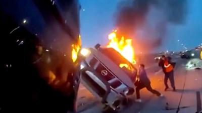 Flames pour out of a car tilted at an angle as people stand nearby.