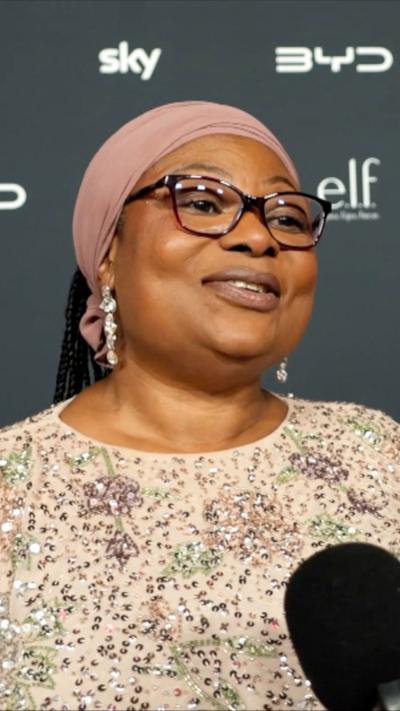 Bemi wears a sparkling beige dress with sequins stands in front of a TikTok Awards backdrop, with a microphone.