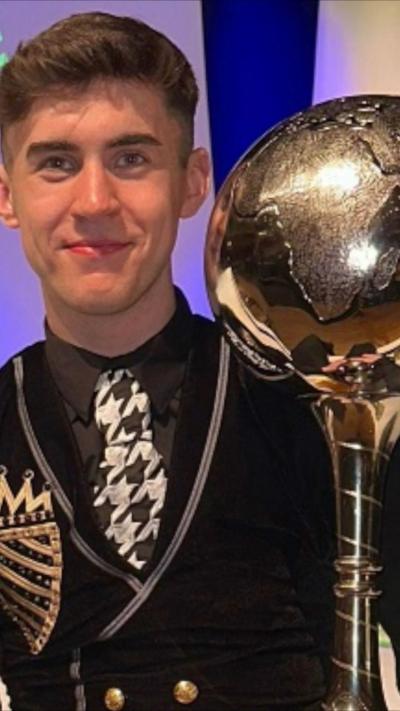 Barry Teague is in a black Irish dancing costume with gold collar trims and a red tie. He is holding a large silver trophy.