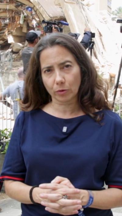 Vanessa Silva in a blue shirt talks in front of a collapsed building