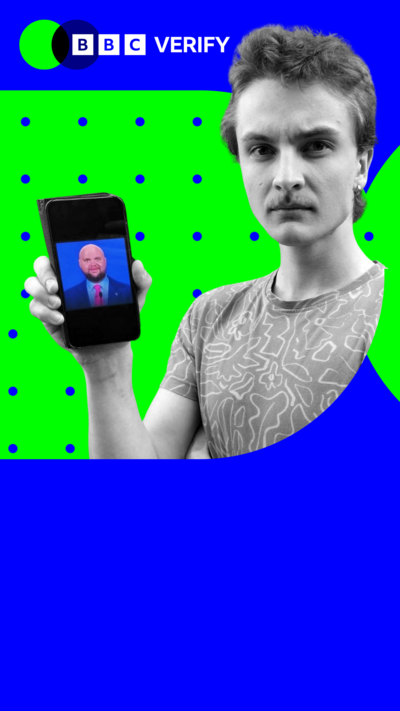A man holds a meme of JD Vance aloft on a phone. In it, Vance is presented as bald with childlike features. 