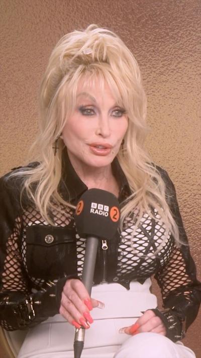 Dolly Parton wearing black mesh shirt speaks into a BBC microphone during sit-down interview