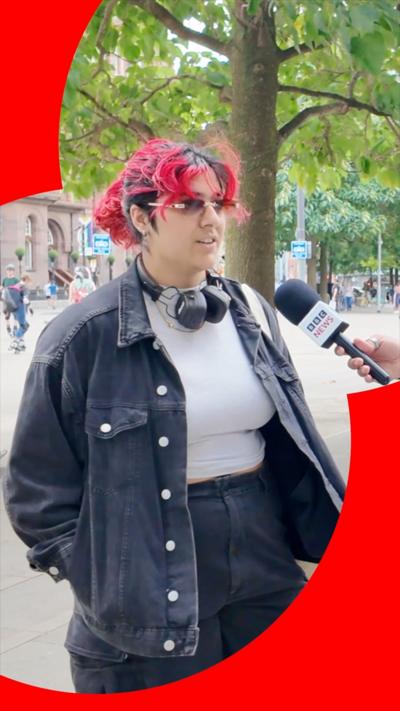 A woman with partially red hair is being interviewed by the BBC - she is wearing dark glasses, a white top, a dark jacket, and dark trousers and is standing outside