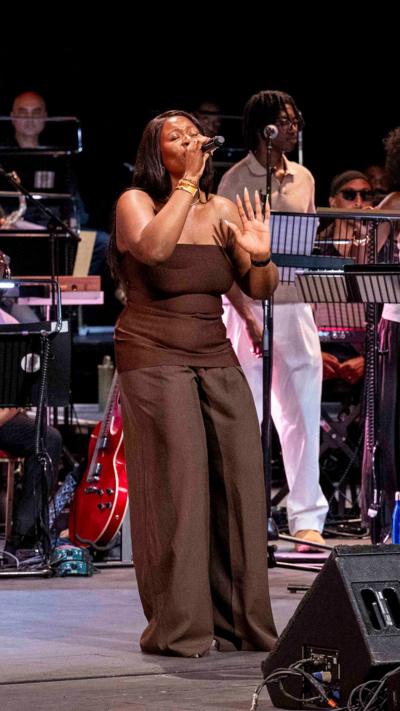 A woman in a brown strapless top and wide-leg pants sings passionately into a microphone on stage, hand raised, with orchestra musicians visible behind her