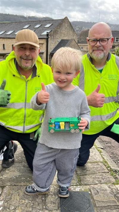 A young boy wearing a grey sweatshirt and holding a green toy truck stands between two waste crew staff, who are also giving the thumbs up. The men are wearing yellow high-visibility jackets