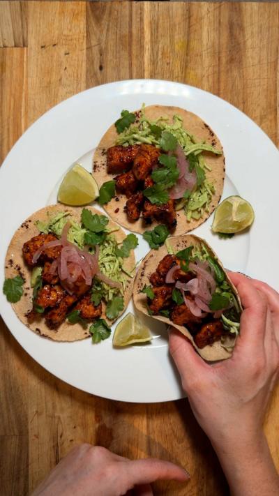 Three tacos on a plate filled with spicy tofu on a wooden surface with a hand reaching in