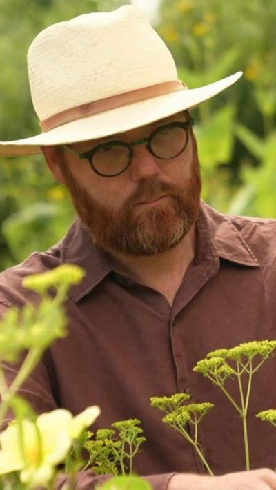 A gardener with a ginger beard and black glasses, wearing a brown shirt and panama hat