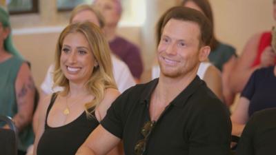 Stacey and Joe, sitting down in an audience, smiling at the stage. Stacey wears a black strapped top, Joe wears a black shirt.