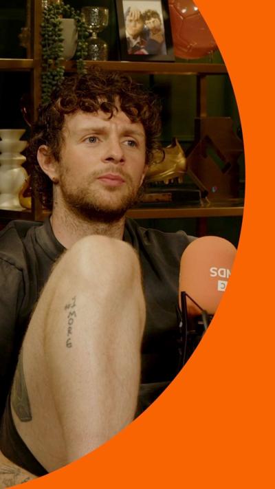 Tom Grennan sits with his knees up, on brown leather sofas in a comfortable studio.