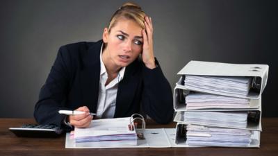 A person holds a pen above a full file of papers with their other hand on their head while looking at a stack of four full files