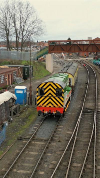 A green shunter train with black and yellow diagonal stripes on the back travelling under a footbridge
