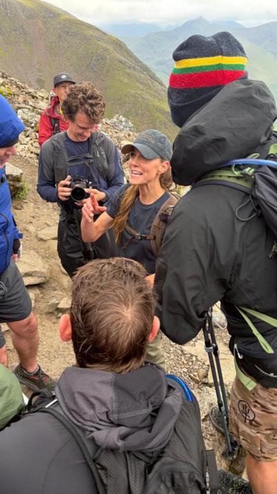 A woman standing in the middle of a crowd pointing animatedly. The crowd are on top of a mountain. The woman is wearing a blue baseball hat. The three men also in shot are wearing black waterproof coats. 