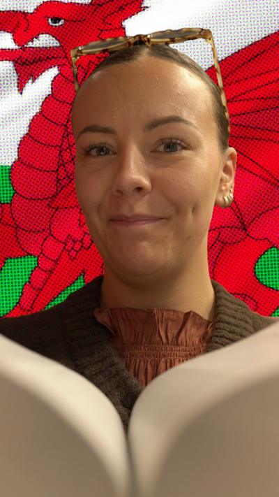 Tink Llywellyn, who has her hair tied back and is wearing tortoiseshell glasses on her head, holding a book close to the camera, superimposed on a flag of Wales