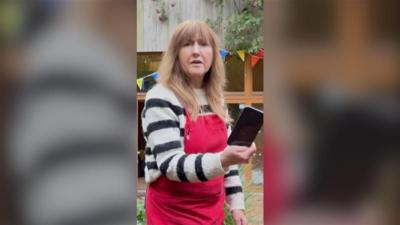 A woman outside with red apron holding a black phone.