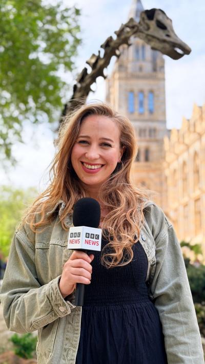 BBC Journalist Sophia Herod, smiling with a microphone outside London’s Natural History museum on a sunny day with trees and a dinosaur sculpture behind her