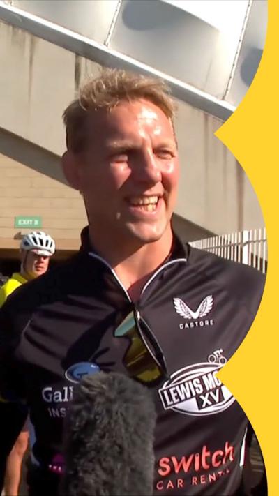 Lewis Moody, a man with short blonde hair, stands looking into the sun, smiling, dressed in cycling gear