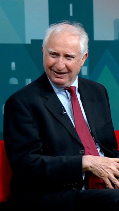A man with white hair and wearing a dark suit, light shirt and red tie is smiling