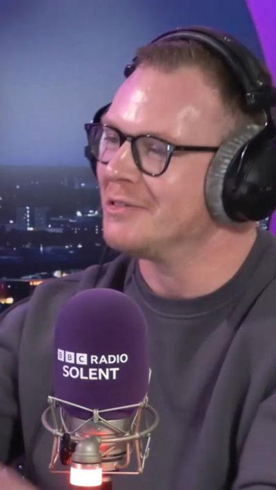 Man wearing glasses and headphones speaking into purple microphone.