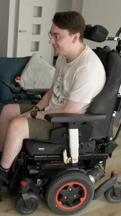 A man with brown hair and glasses in a wheelchair. He is wearing a white t-shirt and green shorts