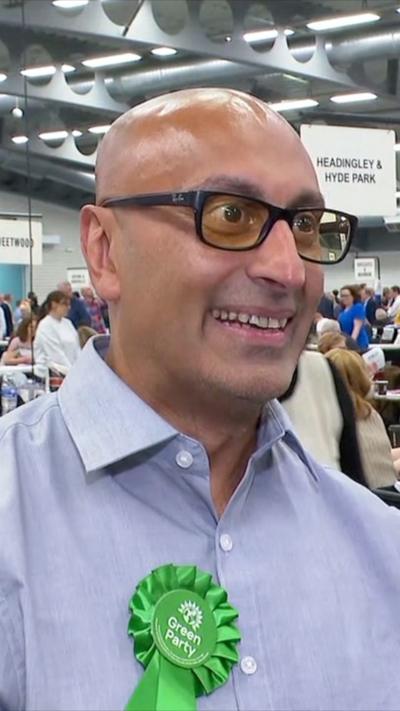 A bald man wearing tinted yellow glasses and a blue shirt wears a green rosette. He is smiling